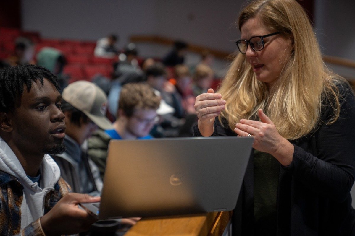An instructor gestures while helping a student working at a laptop in a crowded classroom.