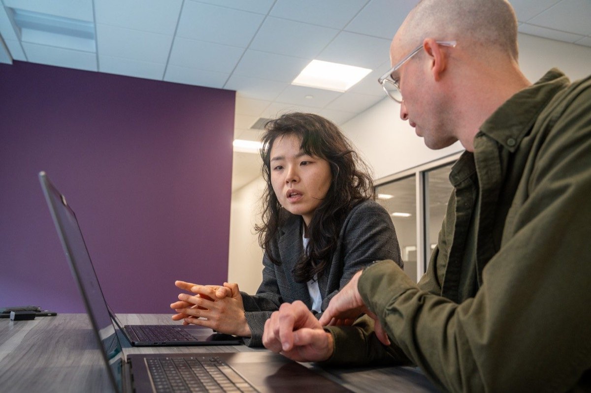 Two colleagues confer over laptop computers in a university workspace.